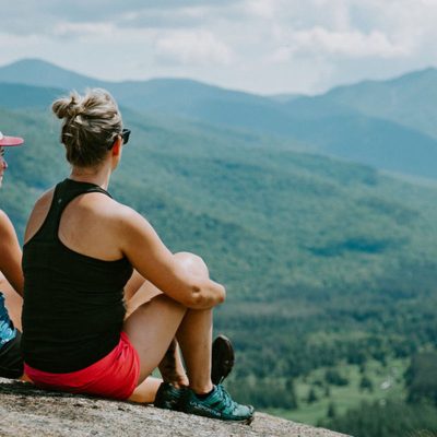 Two women sit on top of a mountain on a summer day looking out at a landscape of the Adirondacks.