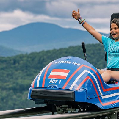 A young girl puts her arms in the air cheering as she rides the Cliffside Coaster. Its summer and the backdrop is the Adirondack Mountains.