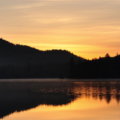 Sunset view of a lake with mountains in the background. It is summer and the mountains are casting a shadow over the lake.