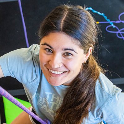A young female climbing up the Hi Notch wall, smiling into the camera above her with hand holds and rope and the floor around her and in the background.