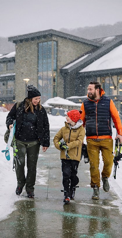 A family of three walks away from the mountain pass lodge holding skis on a snowy winter day.