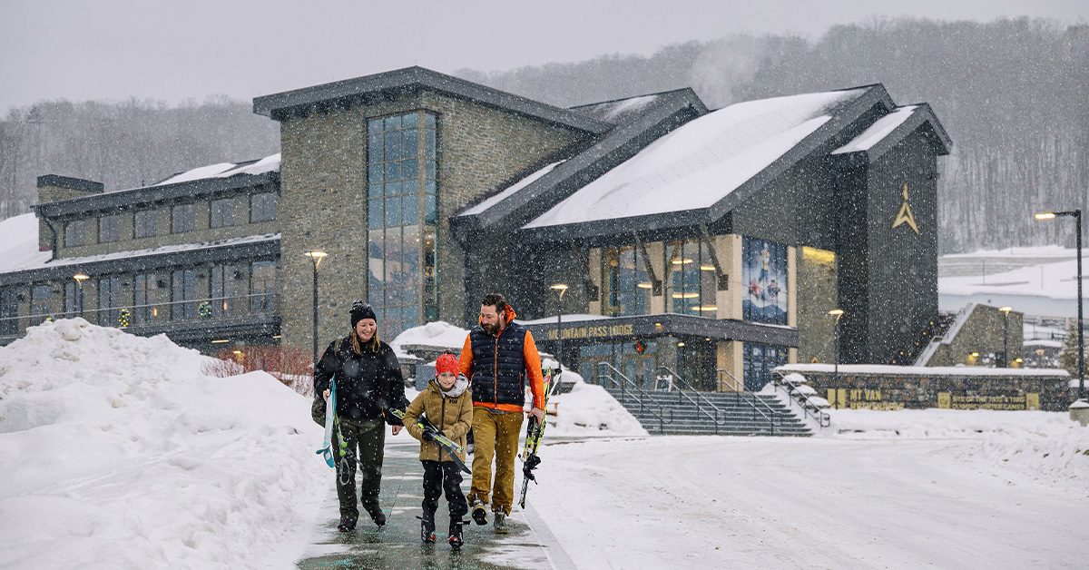 A family of three walks away from the mountain pass lodge holding skis on a snowy winter day.
